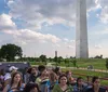 A double-decker tour bus in Washington DC passes by colorful flowers and the iconic dome of the US Capitol under a partly cloudy sky