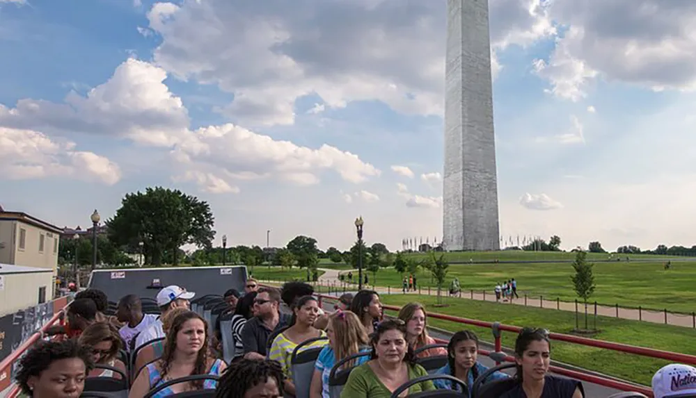 A group of people is seated on an open-top tour bus in front of the Washington Monument on a sunny day