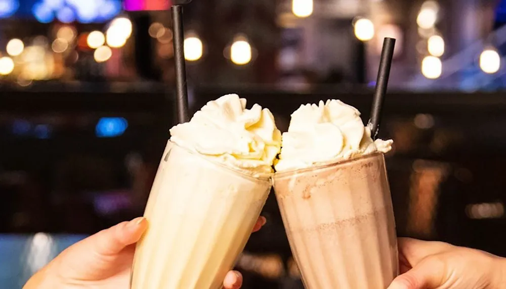 Two hands are holding and clinking together glasses of milkshakes topped with whipped cream