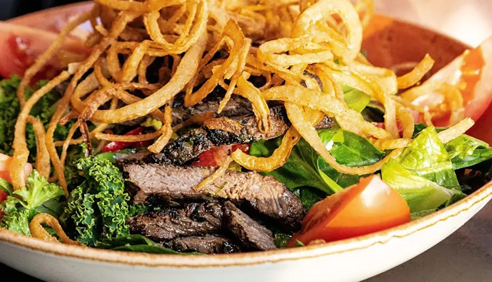 A salad topped with grilled beef slices and crispy fried onions is garnished with fresh greens and tomatoes