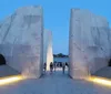 A group of people walks through a large stone monument with illuminated pathways at twilight