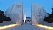 A group of people walks through a large stone monument with illuminated pathways at twilight.