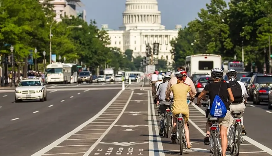 A group of cyclists rides down a designated bike lane towards the United States Capitol building on a busy street with vehicles.