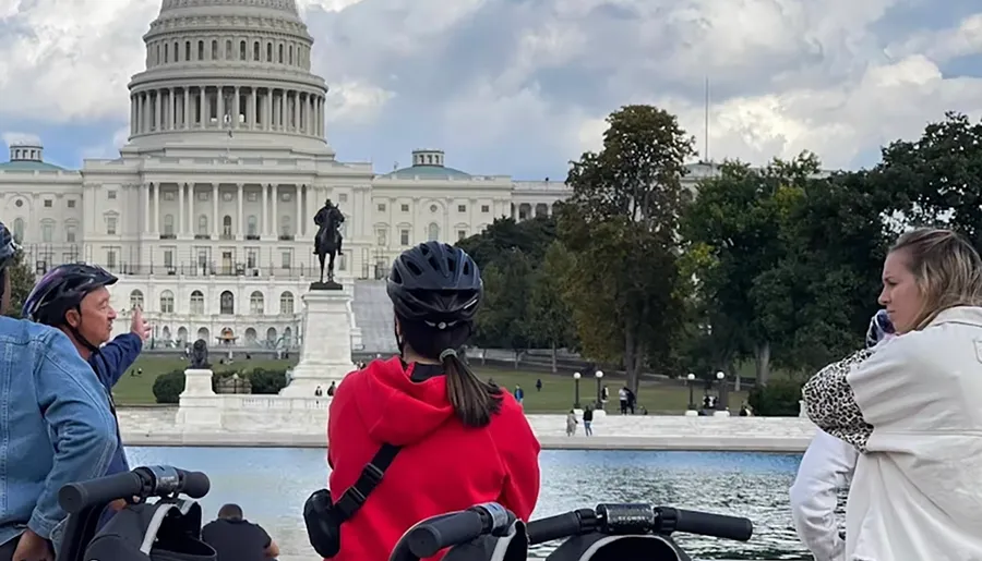 People wearing helmets are gathered in front of the U.S. Capitol building, with a statue and reflecting pool in the foreground.