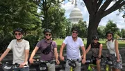 A group of five people wearing helmets pose with bikes in front of the U.S. Capitol building on a sunny day.