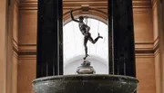 The image depicts a bronze statue of Mercury atop a pedestal within an interior space with tall columns and a skylight.