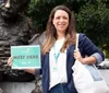 A woman is smiling and holding a Meet Here sign in front of a statue while carrying a tote bag