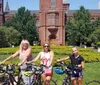 Three people with bicycles pose in front of the Smithsonian Institution Building surrounded by greenery and flowers