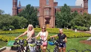 Three people with bicycles pose in front of the Smithsonian Institution Building, surrounded by greenery and flowers.