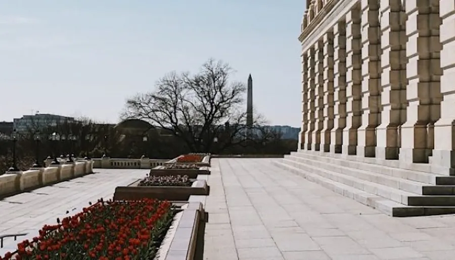 The image shows a view of a stone building with red flowerbeds in the foreground and the Washington Monument visible in the background.