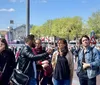 A group of people is gathered outdoors on a sunny day listening to a man in a leather jacket