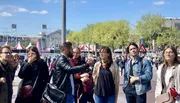 A group of people is gathered outdoors on a sunny day, listening to a man in a leather jacket.