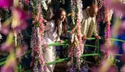 Two people are standing on a bridge adorned with colorful flowers, smiling and looking down.