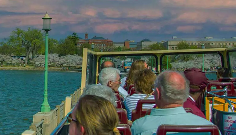 A group of people is riding on an open-top tour bus along a scenic waterfront lined with blooming trees