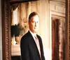 The image depicts a formal oil painting of a man in a suit within an ornate frame hanging on a wall in a gallery