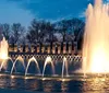 The image depicts a lit fountain with arched streams of water set against a backdrop of trees and a structure with columns at dusk