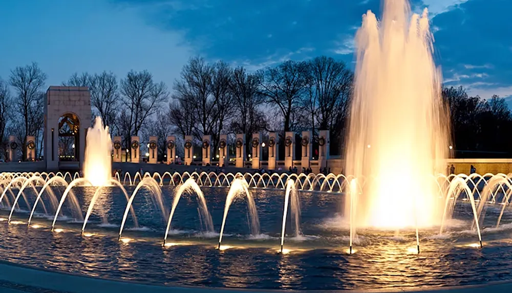 The image depicts a lit fountain with arched streams of water set against a backdrop of trees and a structure with columns at dusk