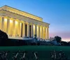 The image depicts a lit fountain with arched streams of water set against a backdrop of trees and a structure with columns at dusk