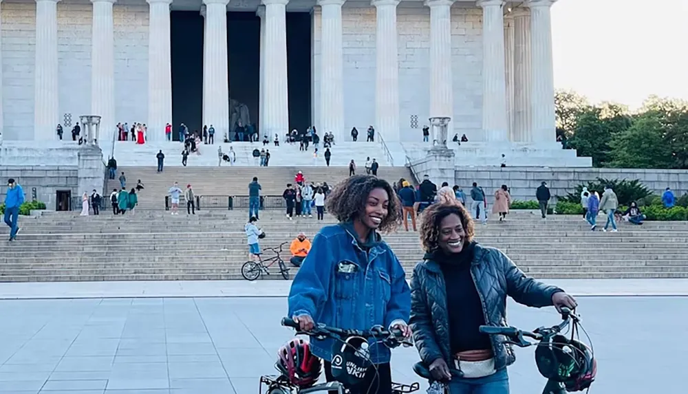 Two people with bicycles smile in front of a large building with columns as others walk up and down the stairs in the background