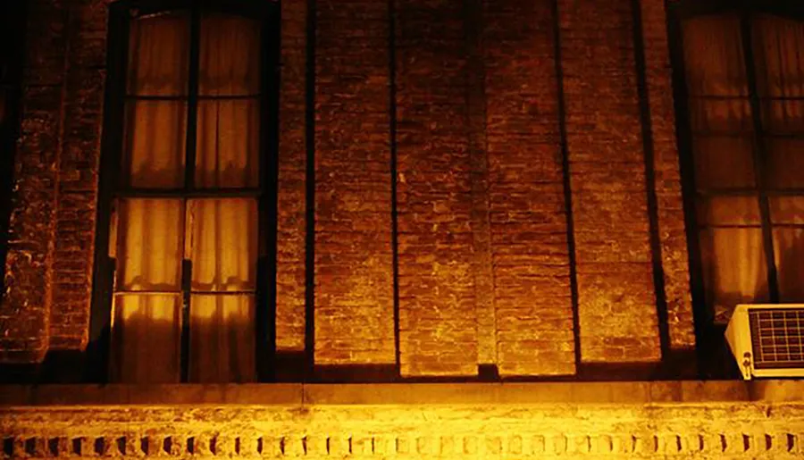 The image shows a warmly lit segment of a brick building at night, featuring tall windows and an air conditioning unit on the right side.