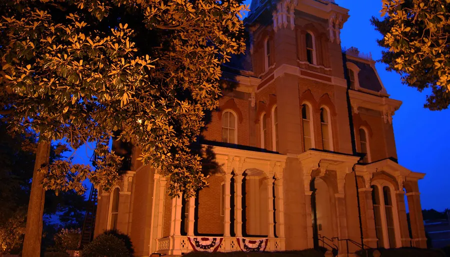 The image depicts a historic building illuminated by warm lights against a twilight sky, surrounded by trees.