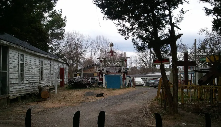 The image shows a rustic and cluttered yard with a weathered wooden structure on the left, assorted materials and sculptures scattered about, and a cat lounging on the driveway.