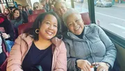 Two women are smiling and sitting together on a bus, with other passengers visible in the background.