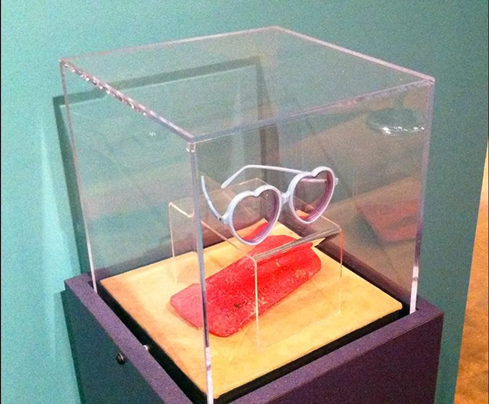 The image shows a pair of glasses with heart-shaped frames placed on a stand inside a clear display case on top of a wooden pedestal