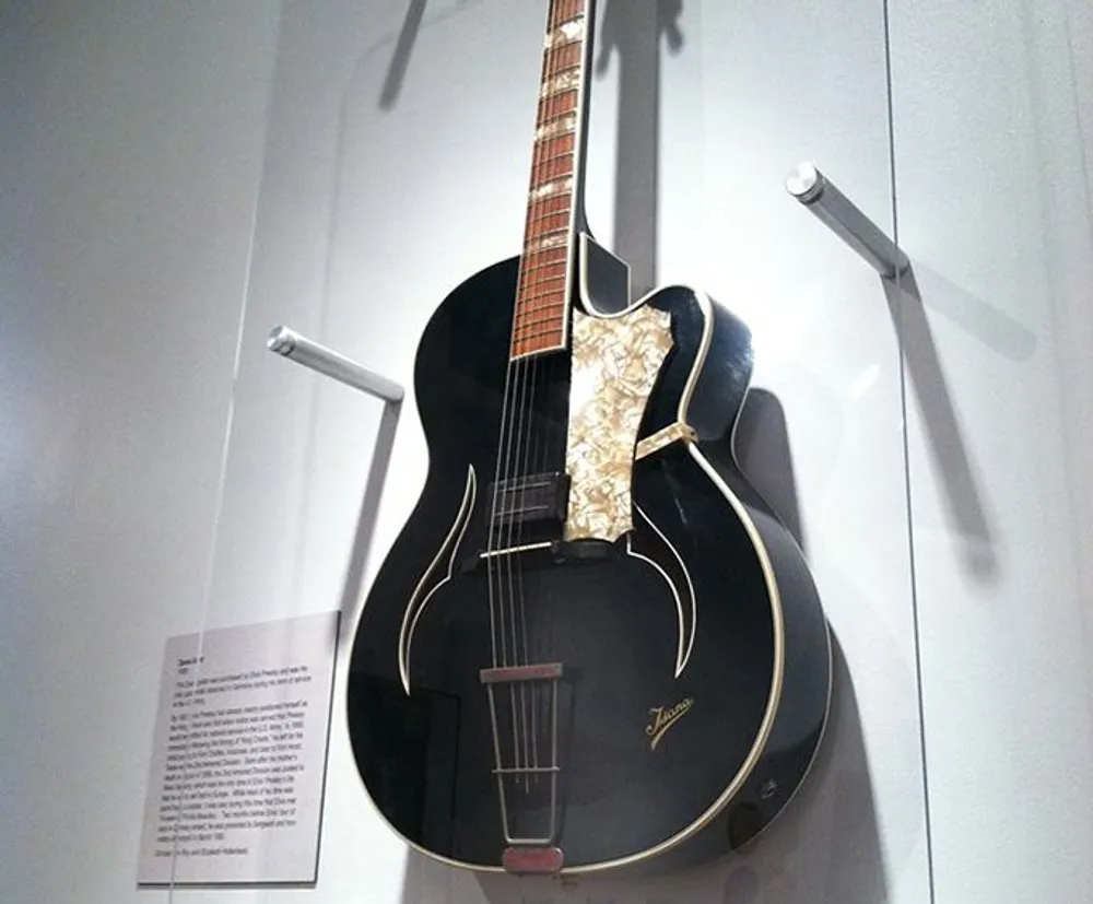 An acoustic guitar with a vintage appearance is displayed on a wall mount featuring a distinctive pickguard and f-holes