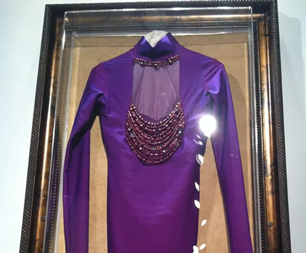 The image features a purple long-sleeve garment with bead embellishments around the neck framed and displayed on a wall giving the impression of a significant or iconic piece of clothing