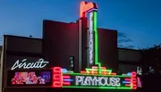 The image shows a brightly lit theater facade with neon signs displaying 