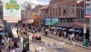 The image shows a lively street scene with people walking among colorful storefronts, neon signs, and outdoor seated areas, reflecting a vibrant urban environment likely known for its music and nightlife.